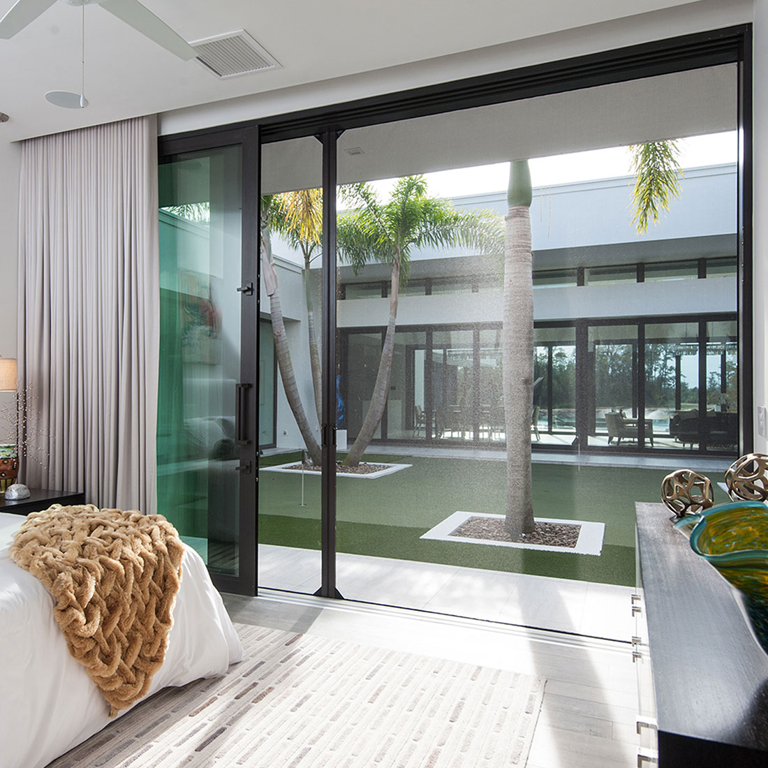 Phantom manual wall screen installed across sliding glass doors in a modern bedroom, with a view of a tropical courtyard and palm trees
