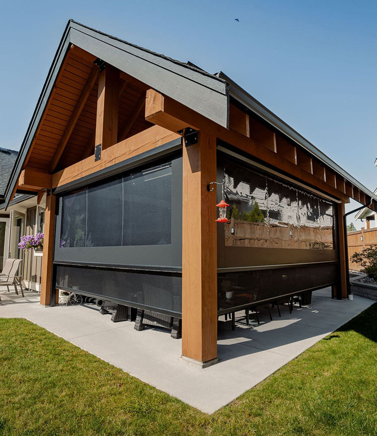 Exterior of a timber-frame backyard structure with large dark retractable screens covering two openings, patio furniture visible underneath, sunny day with blue sky